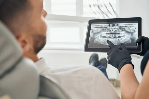 Молодой человек в стоматологическом кресле смотрит на фото зубов на планшете — стоковое фото