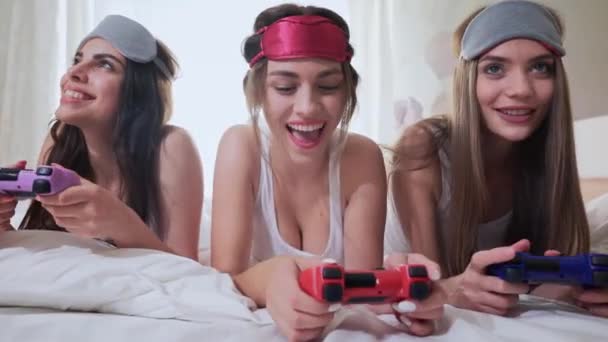 Sonrientes señoritas tumbadas en la cama y jugando videojuegos — Vídeo de stock