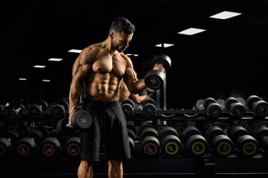 Spor salonunda üzerinde dambıl olan bisepsler yapan tişörtsüz bir adam..