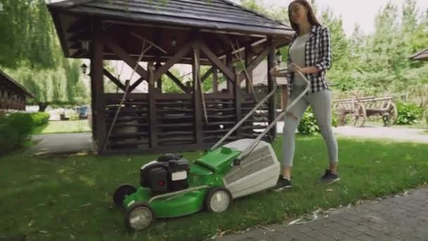 Молодая женщина в клетчатой рубашке стрижет траву газонокосилкой — стоковое видео