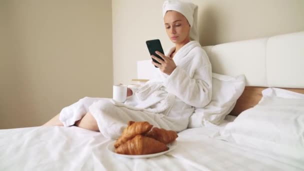 Ragazza utilizzando smartphone, bere caffè sul letto . — Video Stock