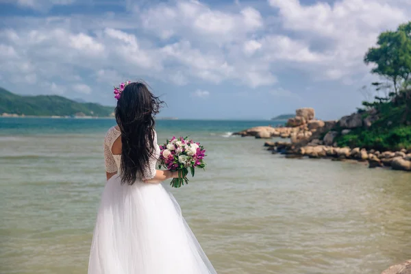 Egy elegáns fehér ruha menyasszony kezében egy csokor, esküvő napján áll a háttérben a tenger. gyönyörű táj — Stock Fotó