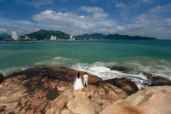 Groom in white suit and the bride in a dress holding each others hands  standing on the background of the sea. beautiful landscape with big stones. concept wedding day — Stock Photo, Image