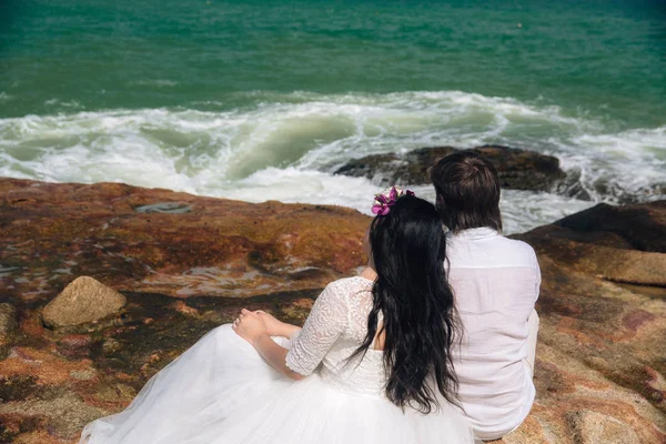 Novio en traje blanco y la novia en un vestido sosteniendo las manos de los demás de pie sobre el fondo del mar. hermoso paisaje con grandes piedras. concepto boda día — Foto de Stock