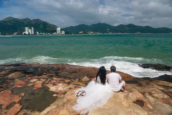 Groom in white suit and the bride in a dress holding each others hands  standing on the background of the sea. beautiful landscape with big stones. concept wedding day — Stock Photo, Image