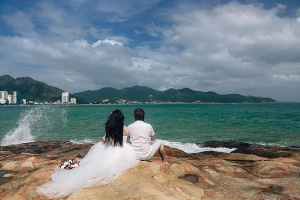 Sposo in abito bianco e la sposa in un abito che si tiene per mano in piedi sullo sfondo del mare. bellissimo paesaggio con grandi pietre. concetto giorno del matrimonio — Foto Stock