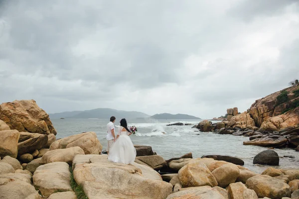 Novio en traje blanco y la novia en un vestido sosteniendo las manos de los demás de pie sobre el fondo del mar. hermoso paisaje con grandes piedras. concepto boda día —  Fotos de Stock