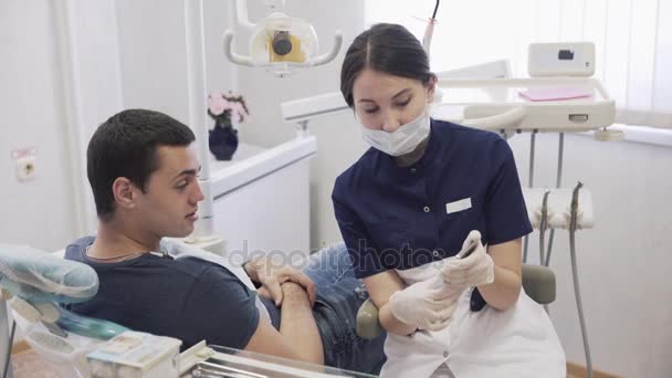 Hermosa mujer dentista mensajes de texto o chat en el teléfono inteligente en guantes estériles y máscara médica y mostrar el teléfono al paciente masculino — Vídeos de Stock