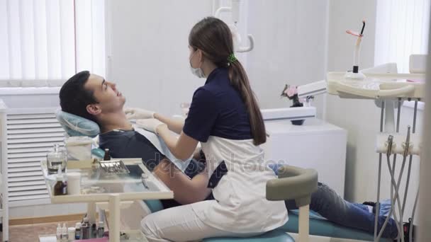 Bela dentista feminina prepara o paciente para os procedimentos na clínica odontológica, coloca no peito pano estéril. Conceito de vida quente — Vídeo de Stock