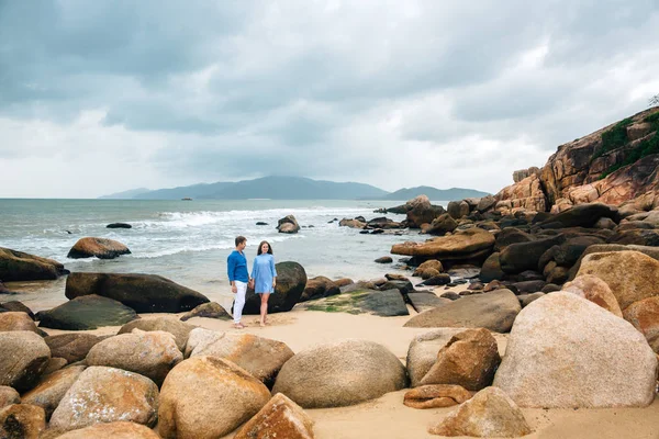 Jovem casal apaixonado em camisa azul de mãos dadas na praia e olhando para o mar. fundo pedras grandes. Conceito de família — Fotografia de Stock