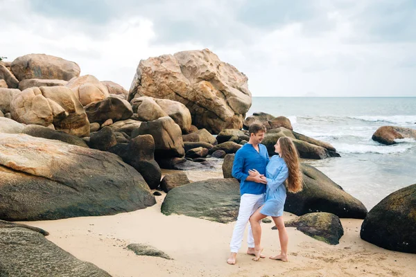 Giovane coppia innamorata in camicia blu che si tiene per mano sulla spiaggia e si guarda. Mare grandi pietre sfondo. Concetto di famiglia — Foto Stock
