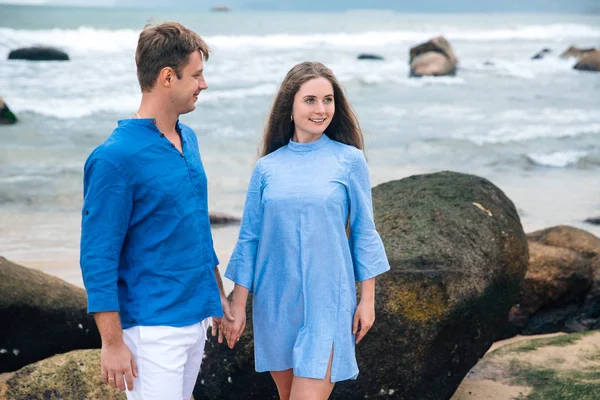 Joven pareja enamorada de camisa azul cogida de la mano en la playa y mirando al mar. piedras grandes fondo. Concepto de familia — Foto de Stock