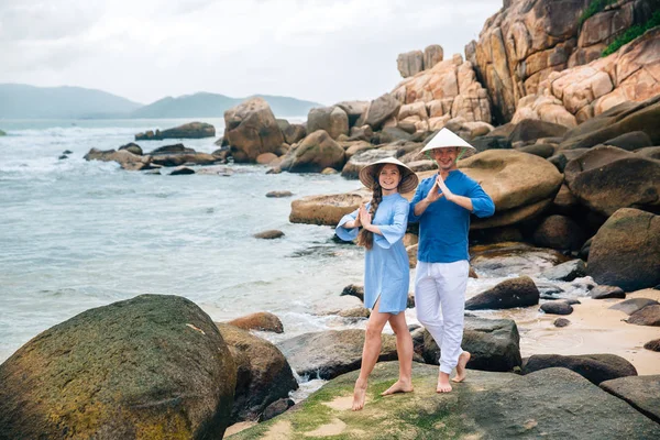 Pareja joven enamorada, camisa azul vestida y sombreros vietnamitas en pose de yoga, tomados de la mano en la playa, mirando al mar. piedras grandes fondo. Concepto de familia — Foto de Stock