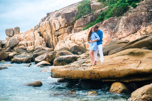 Giovane coppia innamorata in camicia blu tenendosi per mano sulla spiaggia e guardando il mare. grande sfondo di pietre. Concetto di famiglia — Foto Stock