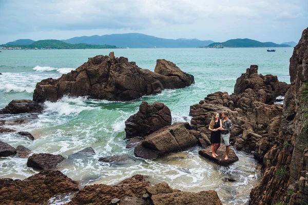 Jovem casal apaixonado em camisa preta de mãos dadas na praia e olhando para o mar. fundo pedras grandes. Conceito de família — Fotografia de Stock