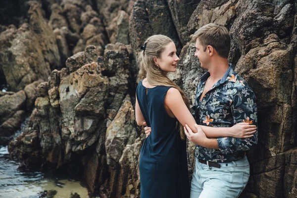 Giovane coppia innamorata in camicia nera che si tiene per mano sulla spiaggia e guarda il mare. grande sfondo di pietre. Concetto di famiglia — Foto Stock