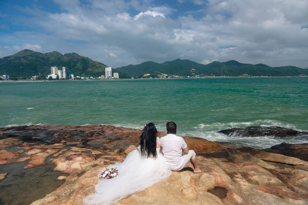 Groom in white suit and the bride in a dress holding each others hands  standing on the background of the sea. beautiful landscape with big stones. concept wedding day — Stock Photo, Image