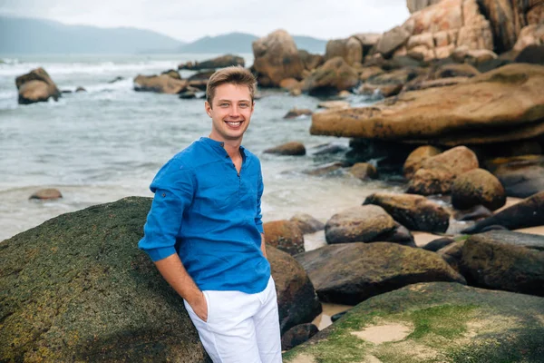 Beau jeune homme blond portant une chemise bleue et un pantalon blanc sur le fond de la mer de grandes pierres souriantes — Photo