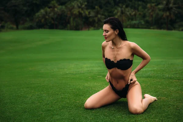 Молода сексуальна і красива брюнетка в чорному купальнику позує на зеленій траві гольф-клубу, тягне труси. Сексуальний портрет моделі з ідеальним тілом. Концепція літнього відпочинку — стокове фото