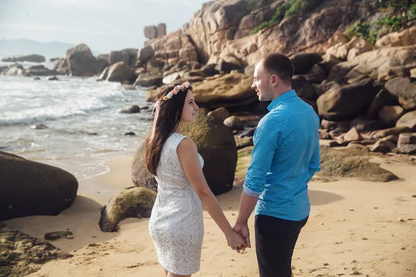 Jovem casal apaixonado abraçando e de mãos dadas em uma praia do mar no verão. O homem de camisa azul e a menina de vestido branco com uma coroa de flores no cabelo — Fotografia de Stock