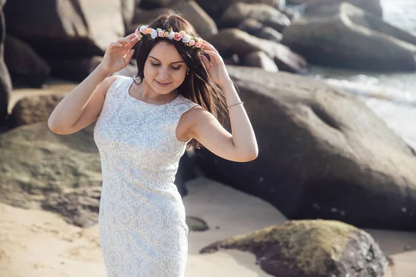 Joven chica hermosa en un vestido blanco en la playa de mar con piedras grandes. Una corona de flores en su cabello. Retrato de belleza de la mujer — Foto de Stock