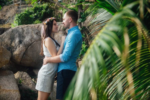 Casal apaixonado abraçando em belo lugar com palmeiras no verão. o homem veste a camisa azul e a menina em um vestido branco com uma grinalda em seu cabelo. conceito de lua de mel — Fotografia de Stock