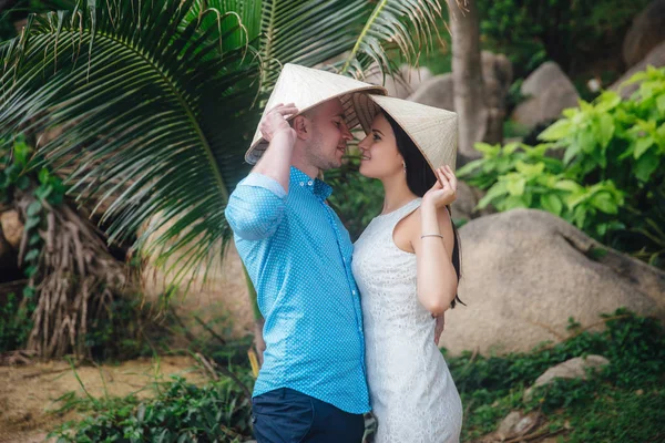 Pareja enamorada abrazándose en un hermoso lugar con palmeras en verano. hombre llevar la camisa azul y la chica en un vestido blanco con sombreros vietnamitas en sus heds. Concepto de luna de miel — Foto de Stock