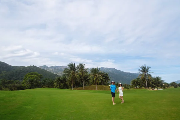 Young beautiful couple in love holding handsand walking at golf course in summer. man wear the blue shirt and the girl in a white dress. Concept of honeymoon — Stock Photo, Image