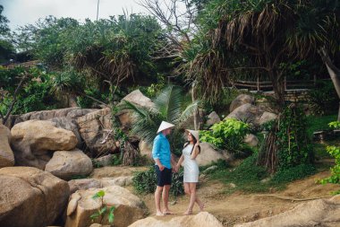 Aşk güzel sarılma içinde çift palmiye ağaçları ile yaz aylarında yerleştirin. adam onların heds üzerinde Vietnamca şapkalı beyaz bir elbise mavi gömlek ve kız giymek. Balayı konsepti