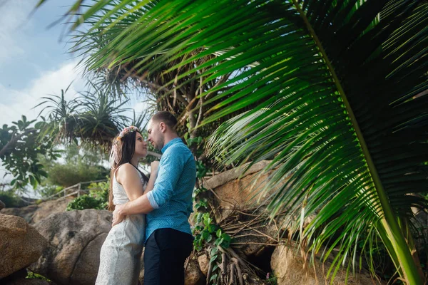 Coppia innamorata che si abbraccia in un bellissimo posto con palme in estate. uomo indossare la camicia blu e la ragazza in un abito bianco con una corona tra i capelli. concetto di luna di miele — Foto Stock