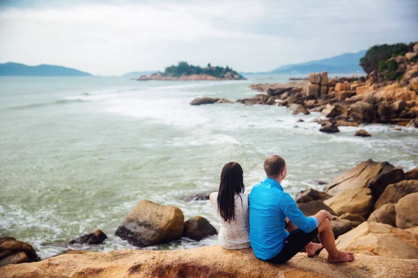 Giovane coppia innamorata seduta a grandi pietre schiena a schiena su una spiaggia di mare in estate. uomo indossare la camicia blu e la ragazza in un abito bianco con una corona tra i capelli — Foto Stock