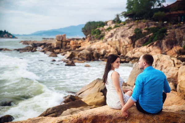 Giovane coppia innamorata seduta a grandi pietre schiena a schiena su una spiaggia di mare in estate. uomo indossare la camicia blu e la ragazza in un abito bianco con una corona tra i capelli — Foto Stock