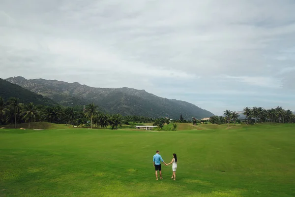 Young beautiful couple in love holding hands and walking at golf course in summer. man wear the blue shirt and the girl in a white dress. Concept of honeymoon — Stock Photo, Image