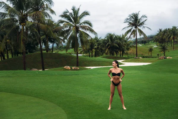 ポーズを回転戦利品黒の水着で若い綺麗でセクシーなブルネットのフィットネス女の子はゴルフ クラブの緑の芝生の上でお尻を示しています。完璧なボディのセクシーなモデルの肖像画。夏の休日の概念 — ストック写真