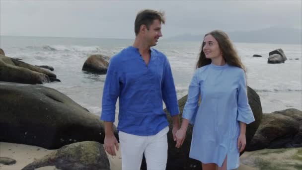 Primer plano joven feliz hermosa pareja de amantes, en ropa azul están tomados de la mano, caminando a lo largo de la orilla del mar, sonriendo, riendo. concepto de una fecha de luna de miel y familia de la felicidad — Vídeo de stock