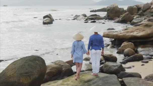 Молодая красивая пара влюбленных в вьетнамских шляпах и голубой одежде, держащихся за руки на берегу моря, смотрящих друг на друга. Концепция медового месяца даты и счастья семьи, путешествия во Вьетнам — стоковое видео