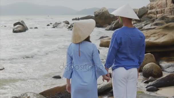 Close up young beautiful couple of lovers in Vietnamese hats and blue clothes holding hands at the beach of sea, looking at each other. Concept of a honeymoon date and happiness family, travel in — Stock Video