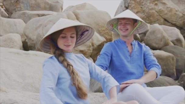 Um cara e uma menina em chapéus vietnamitas sentam-se em rochas na praia em uma pose de ioga, meditam e olham um para o outro. Conceito de uma data de lua de mel e família felicidade, viajar no Vietnã — Vídeo de Stock