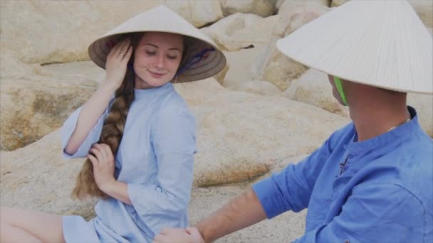Um cara e uma menina em chapéus vietnamitas sentam-se em rochas na praia em uma pose de ioga, meditam e olham um para o outro. Conceito de uma data de lua de mel e família felicidade, viajar no Vietnã — Vídeo de Stock