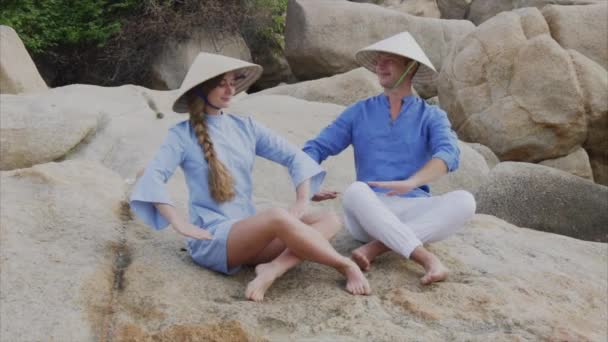 Un chico y una chica con sombreros vietnamitas se sientan en las rocas de la playa en una pose de yoga, meditan y se miran. Concepto de una fecha de luna de miel y la felicidad de la familia, viajar en Vietnam — Vídeo de stock