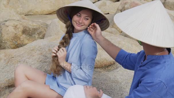 Egy srác és egy lány a vietnami kalap ülni a sziklák a tengerparton, a jóga póz, meditálni, és egymásra nézni. Koncepció egy nászút dátuma és a boldogság család, utazás, Vietnam — Stock videók