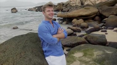 Mavi gömlekli gülümseyen bir sarışın adam büyük taşlar yakınındaki plajda durup ve onun saç düzleştirme