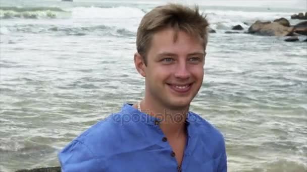 Closeup glimlachend blonde man in een blauw shirt is permanent op het strand in de buurt van grote stenen en zijn haar straightening — Stockvideo