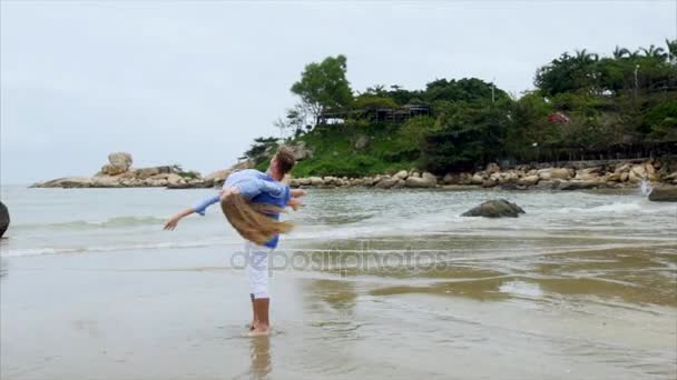 Hebt der Mann das Mädchen in den Arm und umkreist es. junges Paar in blauer Kleidung fliegt auf den Sandstrand — Stockvideo