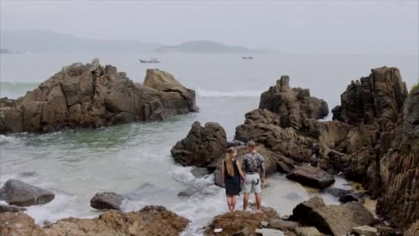 Joven pareja de chicas y hombres están de pie en la playa y tomándose de las manos. Hermosa vista del océano y grandes rocas — Vídeos de Stock