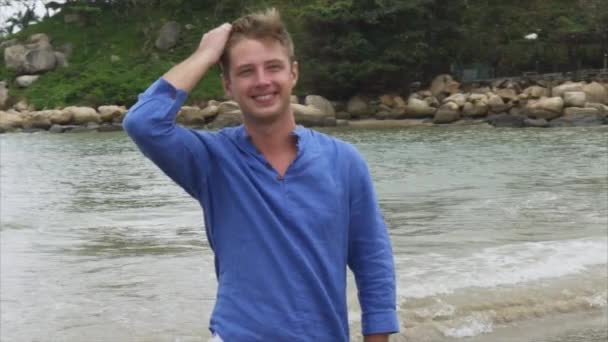 Bliska uśmiechający się blond człowiek w niebieską koszulę jest chodzenie na plaży w pobliżu dużych kamieni i prostowania włosów — Wideo stockowe