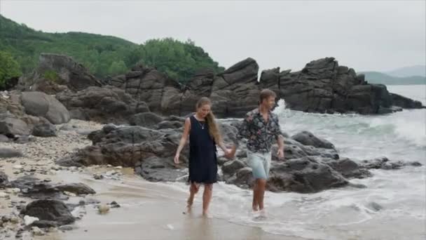 Молодая пара девушка и мужчина прогуливаются по пляжу разговаривая и держась за руки, вид спереди. Красивый вид на океан и большие скалы — стоковое видео