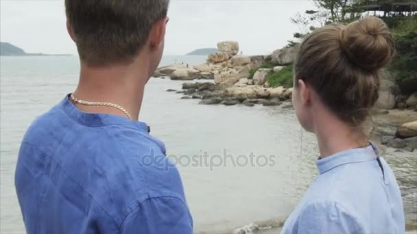 Zblízka, muž ukazuje ruku směrem k obzoru a velké kameny na břehu moře. Mladý pár, drželi se za ruce na břehu oceánu — Stock video