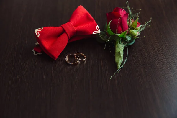Accesorios de boda de novio. Boutonniere rojo, anillos de oro y pajarita sobre fondo marrón —  Fotos de Stock