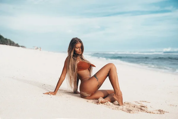 Сексуальна татуйована молода дівчина в червоному купальнику позує на пляжі. Красива блондинка з довгим волоссям, що розслабляється в океані. Концепція спортивної моделі, купальник — стокове фото